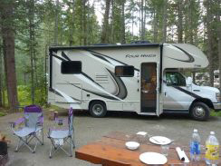 Wohnmobil im Wald, davor ein Tisch mit Geschirr und zwei Campingstühlen.