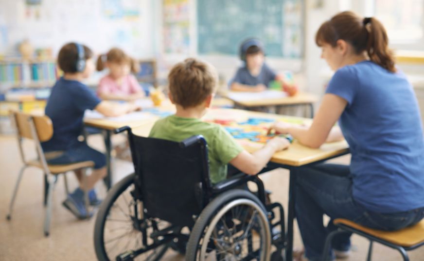 Eine Erzieherin sitzt mit einem Jungen im Rollstuhl an einem Tisch.